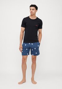 T-shirt noir à manches courtes avec le logo rouge "HUGO" sur la poitrine, associé à un short de bain bleu présentant un motif répété du texte "HUGO" en bleu et blanc.