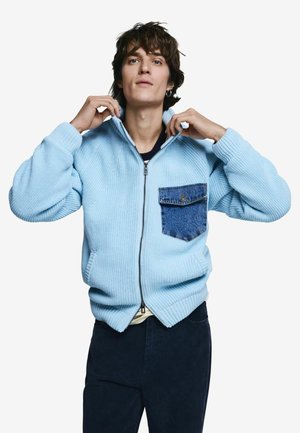 Osoba si upravuje golier svetlomodrého pleteného svetra na zips s modrým denimovým vreckom na hrudi, oblečená v tmavých nohaviciach na jednoduchom pozadí.