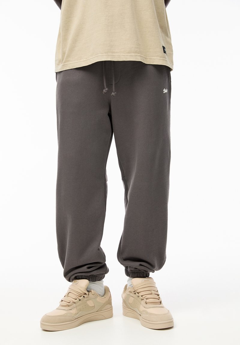 Personne portant un pantalon de survêtement gris foncé avec des poignets élastiques et des baskets beige, debout devant un fond blanc uni.