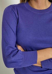 Lila pletena jumper s okroglo izrezom in rebrastimi manšetami. Ima napihnjene rokave in prilagojeno telo. Gladka tekstura in enotna barva.