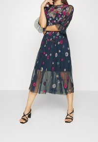 Mörkblå tyllklänning med blommig brodyr i rosa och silver. Har ett genomskinligt liv och en midilång kjol. Matchas med svarta sandaletter.