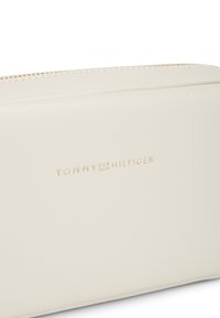 Κρεμ θήκη Tommy Hilfiger με χρυστό φερμουάρ και μικρό ανάγλυφο λογότυπο της μάρκας στο κέντρο της μπροστινής επιφάνειας.