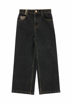Jeune fille aux longs cheveux blonds porte un chemisier imprimé léopard avec un grand col, un pantalon noir à jambes larges et des baskets rouges, debout de côté.