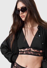 Top bikini nero con scollo a V e fascia leopardata; la modella indossa una camicia nera con bottoni e occhiali da sole squadrati. Capelli lunghi e ondeggianti di colore castano.