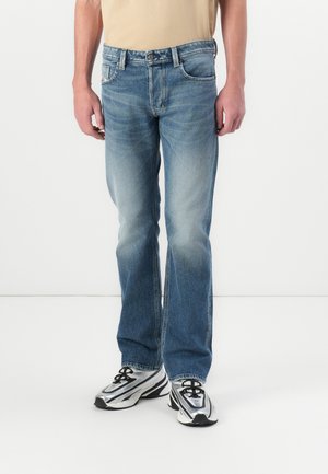Person trägt ausgewaschene blaue Straight-Leg-Jeans, beige Hemd in die Hose gesteckt und silber-schwarze Sportschuhe, steht auf weißem Boden.
