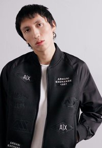 Giubbotto bomber nero con texture a rete e maniche in tessuto solido. Presenta i loghi: "ARMBANI EXCHANGE 1991" e "AIX" con accenti bianchi e neri.