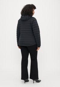 Schwarze Pufferjacke mit Kapuze, mit horizontalem Steppdesign, weichem Material und elastischen Bündchen. Kombiniert mit ausgestellt geschnittenen schwarzen Hosen und High Heels.