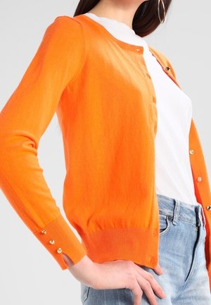 Femme portant un cardigan orange à manches longues boutonné par-dessus une chemise blanche et un jean bleu, debout avec la main sur la hanche.