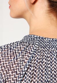 Blusa trasparente con motivo a zigzag nei colori blu navy, bianco e marrone. Caratterizzata da un colletto arricciato e una texture di tessuto fluente.