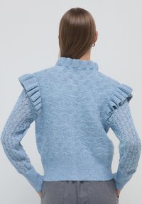 Donna con capelli castani lisci che indossa un maglione lavorato a maglia di un blu chiaro con dettagli a rouches sulle spalle e maniche traforate a motivo, in piedi con le mani sui fianchi.