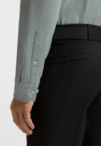 Der Arm eines Mannes trägt ein hell strukturiertes graues Hemd mit geknöpftem Ärmel und liegt neben einer schwarzen Anzughose mit einer Gesäßtasche.