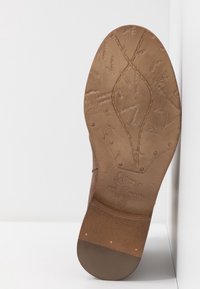 Semelle de botte en cuir marron avec couture décorative, marque en relief et numéro de taille 37 visible sur une surface et un fond blancs.