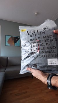 Černá taška vyrobená ze 100% recyklovaného plastu, zabalená v průhledném materiálu s výrazným textem obsahujícím informace o recyklaci a pokyny pro péči.