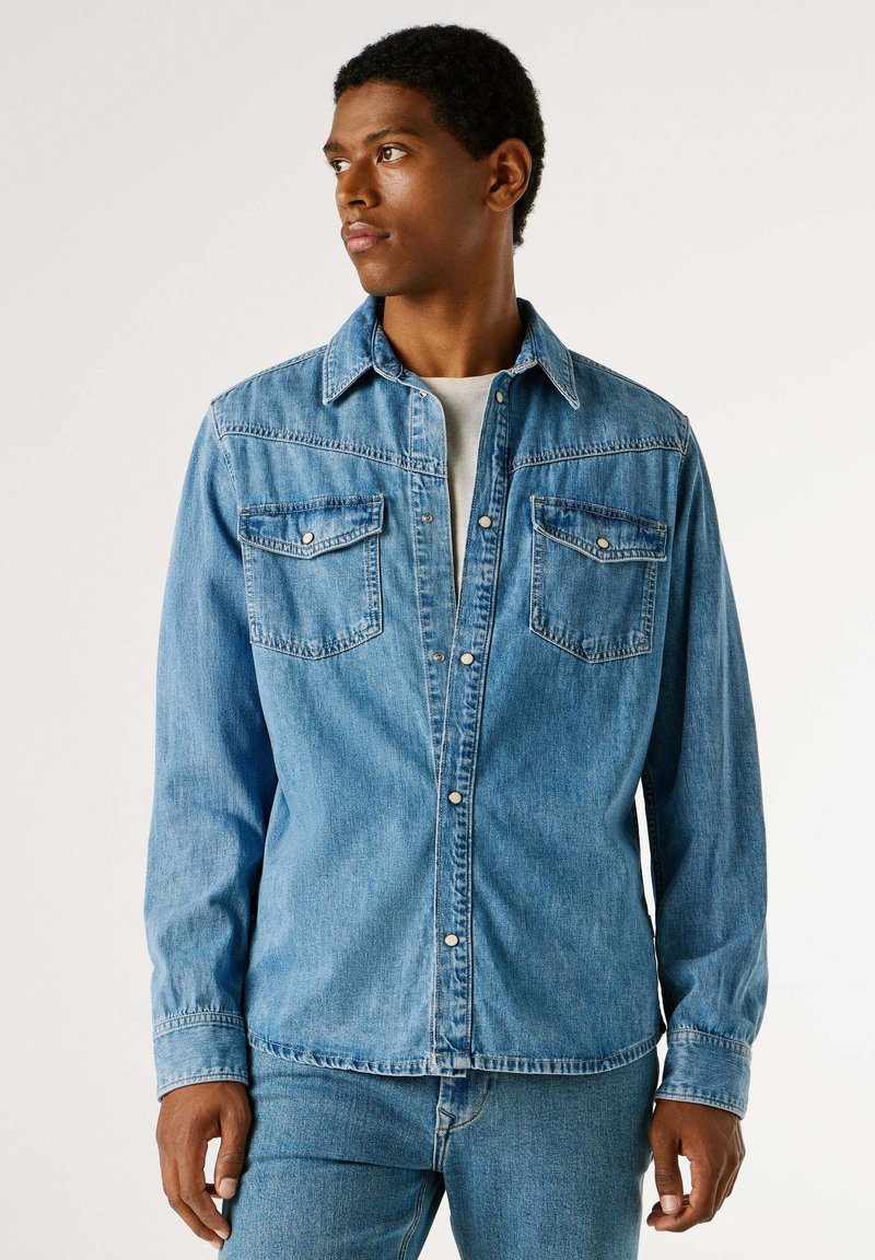 Světle modrá džínová košile se špičatým límcem, dvěma náprsními kapsami a zapínáním na knoflíky. Má dlouhé rukávy a klasický volný střih.