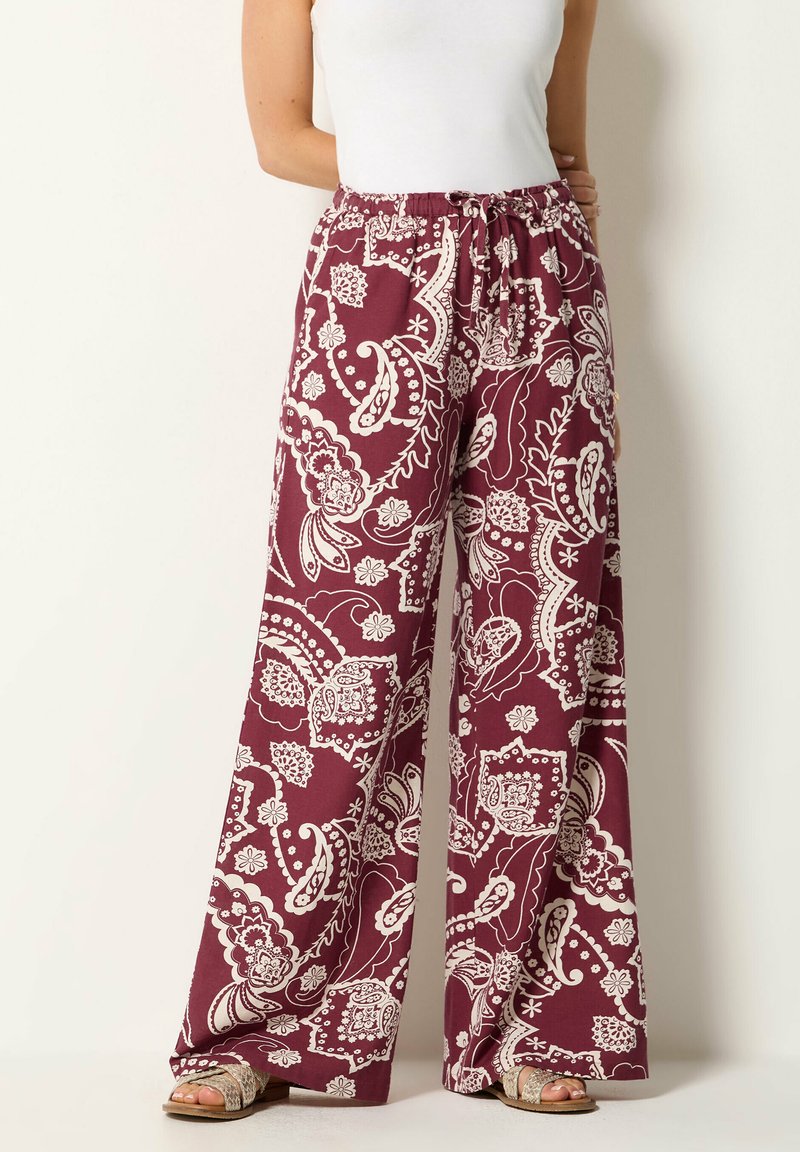 Woman wearing wide-leg maroon pants with white floral paisley print and a white sleeveless top, standing against a plain background.