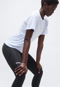 Άτομο που φοράει λευκό κοντομάνικο πουκάμισο και μαύρα κολάν, σκυφτό με τα χέρια στα γόνατα, σε απλό φόντο.