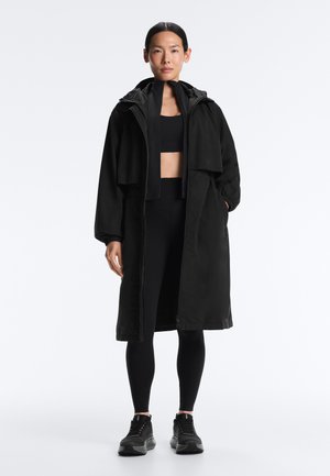 Žena stoji, nosi crni dugi kapuljačasti kaput preko crnog sportskog grudnjaka, crne tajice i crne tenisice, na jednostavnoj svijetloj pozadini.