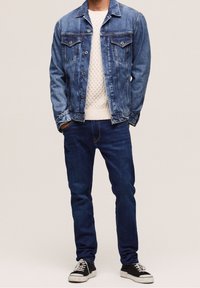 Denim jacket in medium blue, featuring button closures and chest pockets, paired with a white knitted sweater and dark blue jeans, completed with black sneakers.