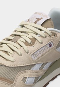 Prim-plan al unei adidași Reebok bej și albi, arătând șireturile texturate, panourile din piele întoarsă, logo-ul Union Jack și limba încălțămintei cu design de frunză de arțar.