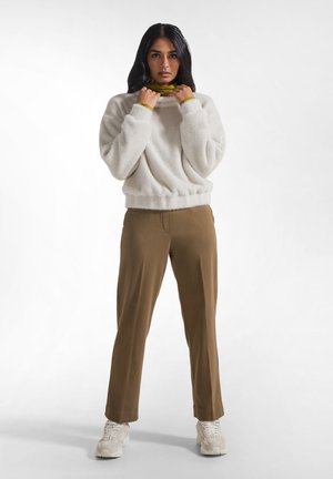 Mujer de pie con un suéter blanco y esponjoso sobre un jersey de cuello alto verde, pantalones marrones y zapatillas blancas sobre un fondo liso.