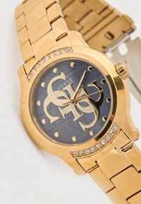 Zapestna ura iz zlatega kovinskega materiala z modrim teksturiranim številčnico, ki vključuje velike zlate številke, kristalne poudarke in zapestni pas v obliki členov.