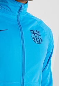 Blaue Nike Zip-Jacke mit FCB-Logo, leichtes Material, hoher Kragen und glatte Textur. Reißverschluss und keine sichtbaren Muster.