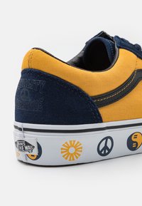 Baskets jaunes et bleu marine avec une tige en toile, des accents en daim, des coutures noires, et une semelle blanche ornée de motifs colorés de paix et de soleil.