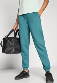 Φόρμες σε τυρκουάζ με χαλαρή εφαρμογή, με ribbed τελειώματα στις μανσέτες, πλαϊνές τσέπες. Μαύρη τσάντα duffel με διπλούς ιμάντες. Μαύρα αθλητικά παπούτσια με υφή από δίχτυ.