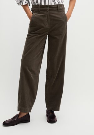 Person wearing dark brown wide-leg pants with pockets and dark brown loafers, hands in pockets, against a plain light background.