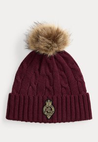 Căciulă tricotată burgundy cu model în cabluri, având un pom-pom din blană bej în vârf și un patch cu logo auriu și negru pe manșetă.