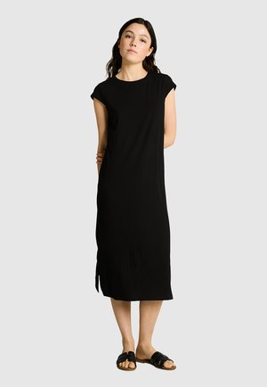 Mujer con vestido midi negro sin mangas con abertura lateral y sandalias planas negras, de pie frente a un fondo liso.