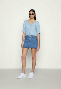 Blusa azul clara de manga curta, com botões e cintura ajustável, combinada com uma saia mini de jeans e sapatilhas brancas. Óculos de sol redondos usados.