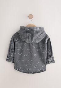 Giacca di jeans grigia con cappuccio, caratterizzata da un motivo ripetuto di faccine sorridenti. Dotata di polsini con bottoni e appesa a una gruccia di legno.