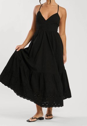 Femme portant une robe noire sans manches avec une broderie œillets à l'ourlet, tenant la jupe, et des sandales plates noires pieds nus.