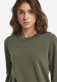 Zielono-oliwkowy sweter z okrągłym dekoltem, długimi rękawami i miękkim, teksturowanym materiałem. Zbliżenie, podkreślające detale przeszycia.