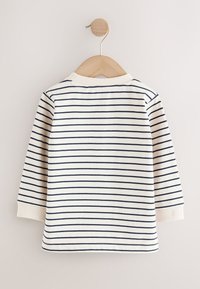 Chemise blanche à manches longues avec des rayures horizontales noires, suspendue sur un cintre en bois contre un mur beige.