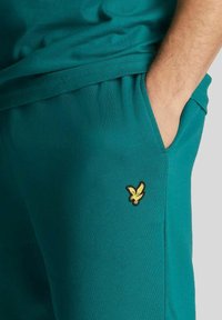 Pantaloni jogger color teal con una texture morbida, dotati di una tasca laterale e di un logo di un'aquila ricamato in giallo sul davanti.