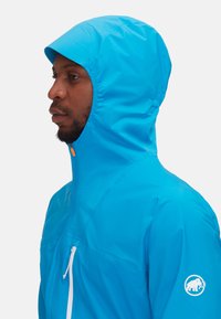 Veste imperméable bleue avec capuche ajustée, fermeture éclair à l'avant et zippes blanches en accent. Logotype sur la manche gauche. Texture lisse.