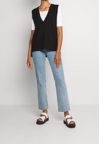 Veste noire en maille avec un col en V, superposée sur un t-shirt blanc à manches courtes. Associée à un jean droit bleu clair et des sandales blanches à brides.