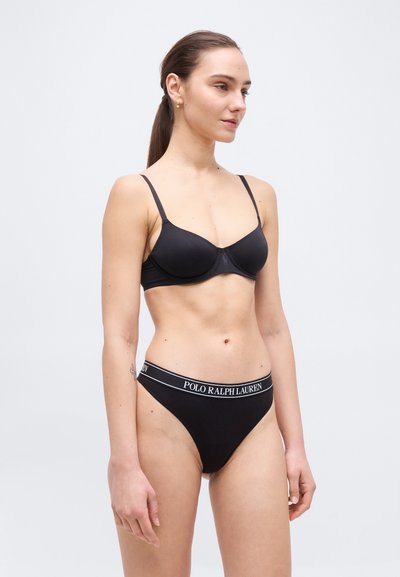 Γυναίκα που φορά μαύρο σουτιέν και εσώρουχο Polo Ralph Lauren, στέκεται μπροστά σε απλό φωτεινό φόντο, στραμμένη ελαφρώς προς τα δεξιά.