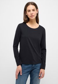 Schwarzes Langarm-T-Shirt aus weichem Baumwollstoff, mit rundem Halsausschnitt und einem kleinen Logo-Detail auf der Brust. Kombiniert mit blauen Denim-Jeans.