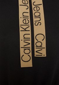 T-shirt noir avec deux étiquettes en tissu beige portant le texte noir "Calvin Klein Jeans" dans une police audacieuse et moderne.