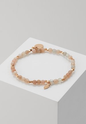 Bracelet délicat à perles avec pierres facettées couleur pêche et gris clair, petites perles intercalaires en or, et un pendentif feuille en or sur un socle blanc.