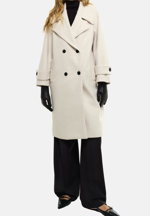 Frau trägt einen beigen zweireihigen Mantel, schwarze Handschuhe, schwarze Hosen mit weitem Bein und schwarze spitze Ballerinas mit Knöchelriemen.