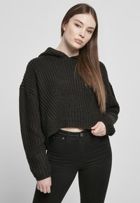 Fekete, kötött cropped kapucnis pulóver, texturált mintával és hosszú ujjakkal, magasderékú fekete farmernadrág felett viselve.