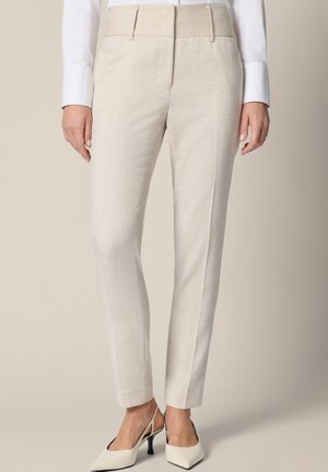 Kobieta nosząca beżowe spodnie o dopasowanym kroju z przednią kantą, połączone z białą koszulą i białymi sandałami na niskim obcasie z paskiem na piętę.