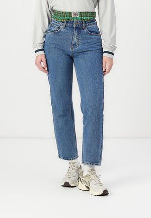 Light blue denim high-waisted jeans with a green plaid elastic waistband, straight leg shape, and contrast stitching. Worn with neutral sneakers.
