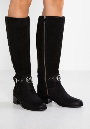 Botas de ante negras hasta la rodilla con cremallera lateral, puntera redonda, tacón grueso y una correa decorativa con un adorno metálico circular.