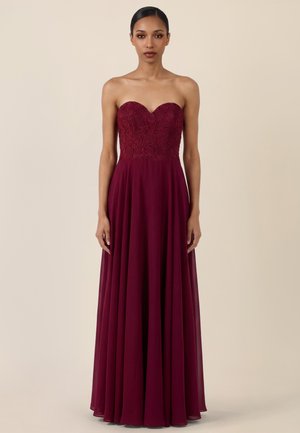 Frau in einem trägerlosen, bodenlangen burgunderroten Kleid mit einem Spitzenoberteil und einem fließenden Chiffonrock, stehend vor einem beigen Hintergrund.