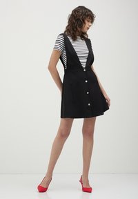 Robe noire avec un décolleté en V profond, des bretelles spaghetti et six grands boutons sur le devant. Portée par-dessus une chemise à rayures blanches et noires. Talons rouges.